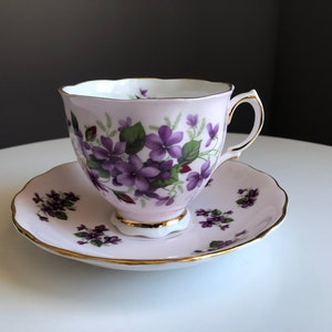 May include: A vintage teacup and saucer set. The light pink porcelain cup and saucer are decorated with purple violets and green leaves. Gold trim accents the edges of the cup and saucer.