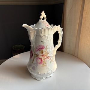 May include: A white porcelain coffee pot featuring an embossed scrollwork pattern and a delicate floral design of pink thistle flowers. The vessel includes a matching lid with a decorative finial, a curved handle, and a scalloped pouring spout.