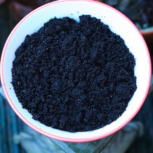 May include: A white bowl with a red rim, filled with dark, granular material. The bowl is round and sits on a wooden surface. The dark material appears to be soil or a similar substance, filling the bowl to the top.