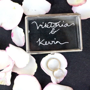 Könnte beinhalten: Eine transparente Glas-Ringbox mit silbernem Rand, die zwei goldene Eheringe enthält. Die Box steht auf einer schwarzen Oberfläche, auf der weiße Rosenblätter verstreut sind. Die Box ist mit den Namen "Viktoria & Kevin" in weißer Kalligraphie beschriftet.