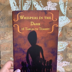 May include: A book cover with a dark purple and orange background. The title is "Whispers in the Dark" with the subtitle "16 Tables to Terrify". The cover features a silhouette of a person standing in front of a group of people.