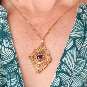 May include: A gold pendant necklace with a diamond-shaped filigree design. The pendant features a large, round, purple gemstone in the center, surrounded by smaller gemstones. The necklace is worn over a teal and white patterned top.