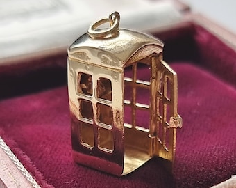 Puede incluir: Un amuleto de color dorado en forma de cabina telefónica británica con la puerta abierta. El amuleto está sentado en una caja de terciopelo rojo.