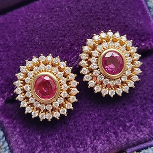 Puede incluir: Un par de pendientes de oro. Cada pendiente presenta una piedra central de color rubí, de forma ovalada, rodeada por un halo de pequeñas piedras preciosas transparentes. Los pendientes están sobre una superficie de terciopelo morado.