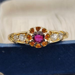 Peut inclure: Une bague en or avec un motif floral. Le centre présente une pierre précieuse rose foncé ronde, entourée d'un sertissage en forme de fleur avec des pétales brun-orangé. Deux petites pierres précieuses claires flanquent la fleur centrale. La bague est dans un écrin bleu.