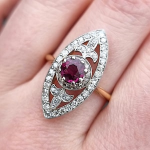 Può includere: Un anello in stile vintage con un grande rubino rosso scuro circondato da un alone di diamanti più piccoli. L'anello è incastonato in un design delicato e filigranato e ha una forma ovale appuntita.