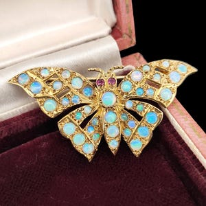 Puede incluir: Broche de mariposa dorada con ópalos y un rubí. Las alas están adornadas con numerosos ópalos de varios tamaños, mostrando tonos azules y blancos. Un pequeño rubí está engastado en el centro del cuerpo de la mariposa.
