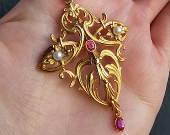 Könnte beinhalten: Ein goldener Anhänger mit einem filigranen, komplizierten Design mit drei roten Edelsteinen und drei weißen Perlen. Der Anhänger ist an einer goldenen Kette befestigt.