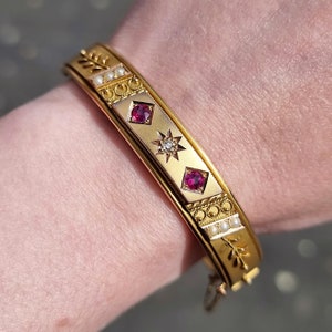 Op de afbeelding: Een gouden armband met een decoratief ontwerp met drie rode edelstenen en een enkele witte edelsteen. De armband is versierd met kleine witte parels.