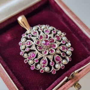 Pode incluir: Um pingente de prata com um design circular que apresenta um grupo de rubis rosa e pérolas brancas. O pingente tem um delicado padrão de filigrana e um laço para prender uma corrente.