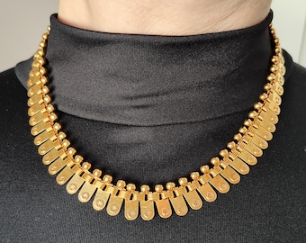 Könnte beinhalten: Eine goldfarbene Halskette mit rechteckigen Anhängern und kleinen kugelförmigen Elementen. Die Kette wird über einem schwarzen Rollkragenpullover getragen. Jeder Anhänger hat ein sternförmiges Muster.