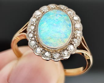 May include: A gold ring featuring an oval opal gemstone with a blue and green iridescence. The opal is surrounded by a halo of small, clear diamonds. The ring has a classic, elegant design.