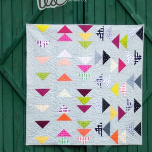 May include: A quilt pattern called "Scrambled Geese" by Jennifer McClanahan. The quilt is made of colorful fabric squares in a variety of patterns and colors, including pink, yellow, green, blue, and white. The quilt is displayed against a green wooden background.