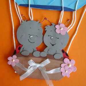 May include: Two gray ladybug characters with red spots and pink flowers are riding in a hot air balloon with white strings. The balloon is blue and white.