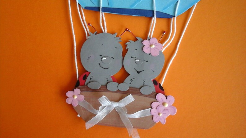 May include: Two gray ladybug characters with red spots and pink flowers are riding in a hot air balloon with white strings. The balloon is blue and white.