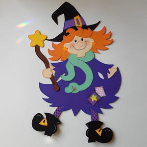 Può includere: Decorazione a tema strega di Halloween. La strega ha i capelli arancioni, un cappello nero con una fascia viola e un vestito viola. Tiene in mano una bacchetta a forma di stella e indossa una sciarpa verde con un serpente. Le scarpe della strega sono nere con dettagli gialli.
