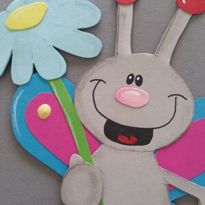 Pu&ograve; includere: Una farfalla di carta grigia con un viso sorridente, antenne rosse e un naso rosa. La farfalla tiene un fiore blu e giallo con uno stelo verde.