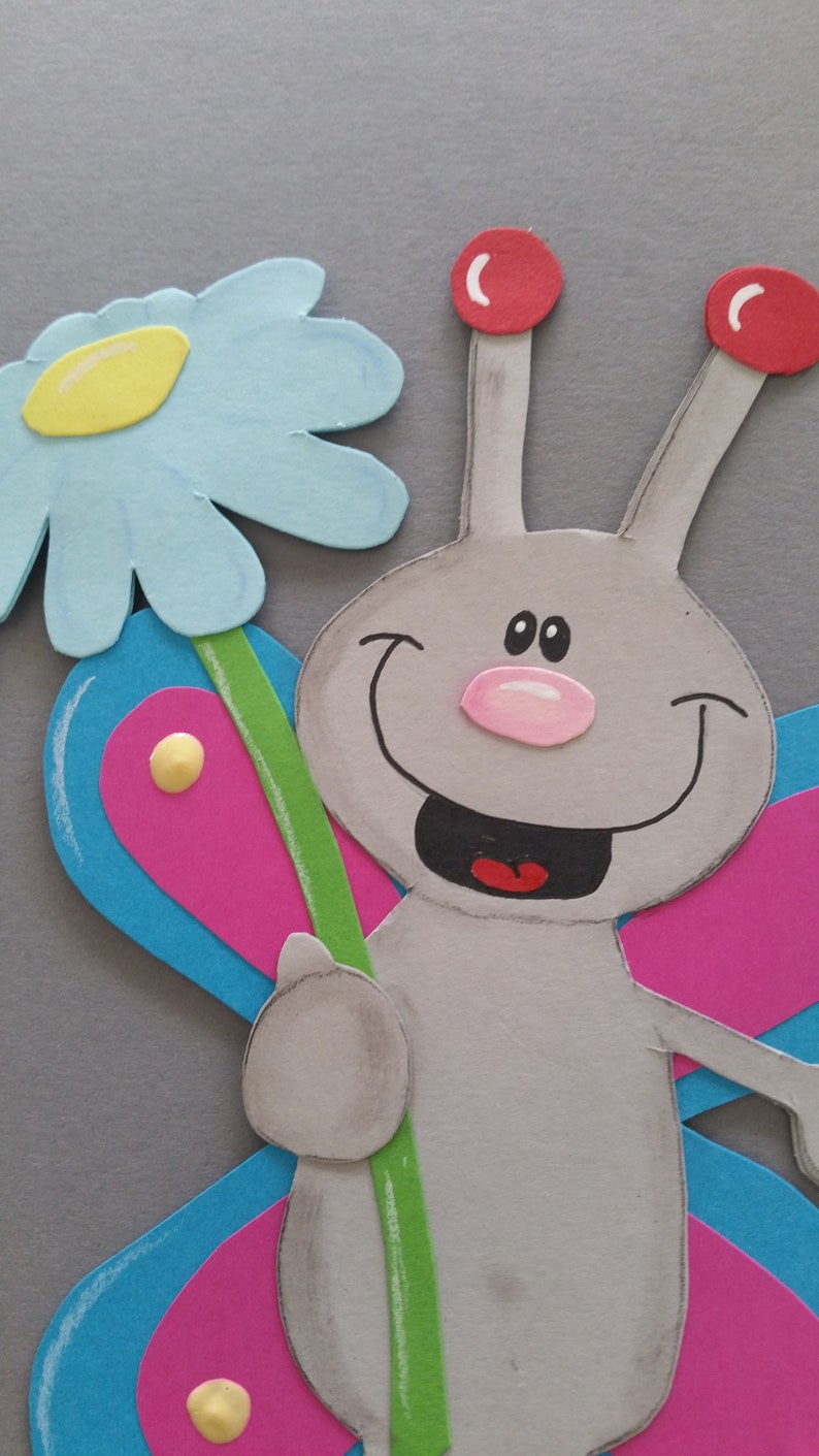 Pu&ograve; includere: Una farfalla di carta grigia con un viso sorridente, antenne rosse e un naso rosa. La farfalla tiene un fiore blu e giallo con uno stelo verde.