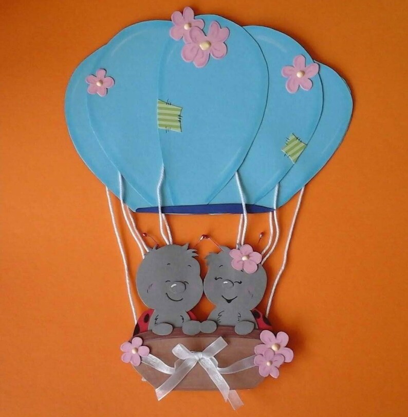 May include: Two gray ladybug characters with pink flowers on their heads are riding in a blue hot air balloon with pink flowers. The balloon is made of paper and has a brown paper basket with a white ribbon.