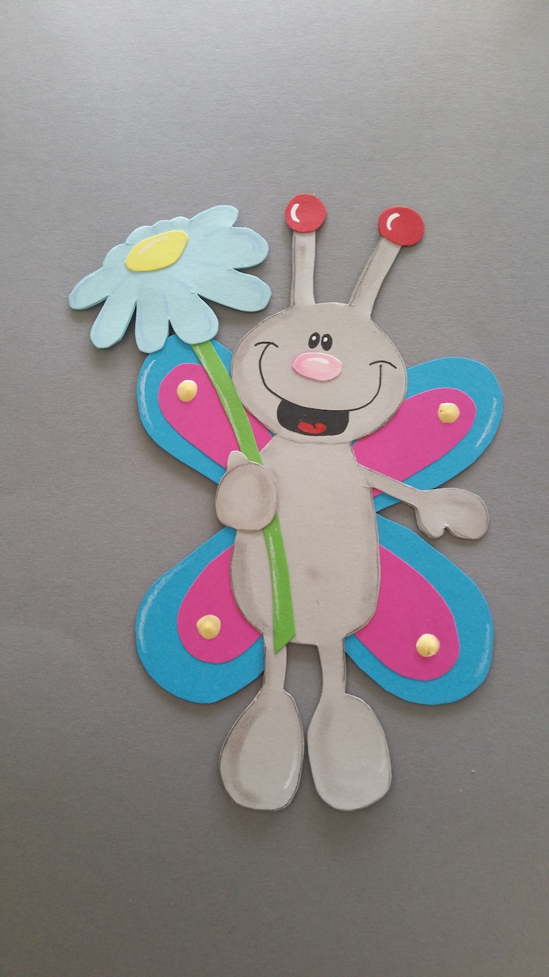 Pu&ograve; includere: Una farfalla grigia con ali rosa e blu tiene un gambo verde con un fiore blu chiaro. La farfalla ha un viso sorridente con antenne rosse e un naso rosa.