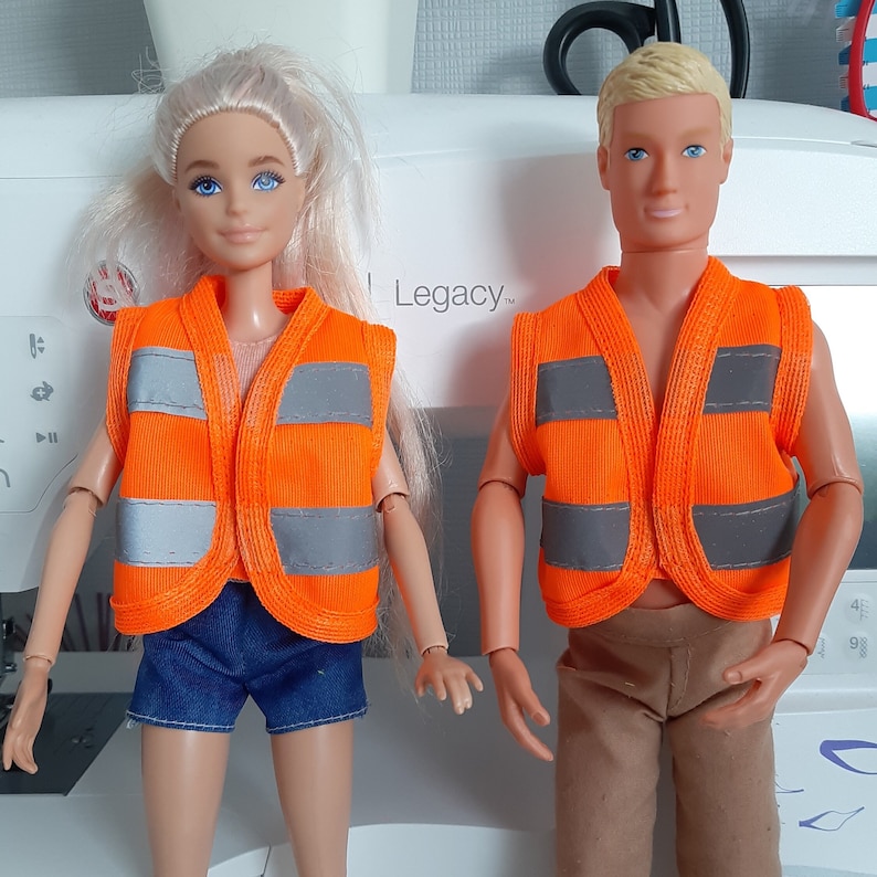 Puede incluir: Dos mu&ntilde;ecas que llevan chalecos de seguridad naranjas con tiras reflectantes. Las mu&ntilde;ecas llevan ropa diferente, una lleva pantalones cortos de mezclilla azul y la otra pantalones marrones. Las mu&ntilde;ecas est&aacute;n de pie frente a una m&aacute;quina de coser blanca con la palabra "Legacy" impresa en ella.