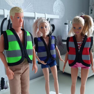 Puede incluir: Tres mu&ntilde;ecas con chalecos de seguridad. Los chalecos son verde, azul y rojo con rayas reflectantes plateadas. Las mu&ntilde;ecas llevan ropa informal.