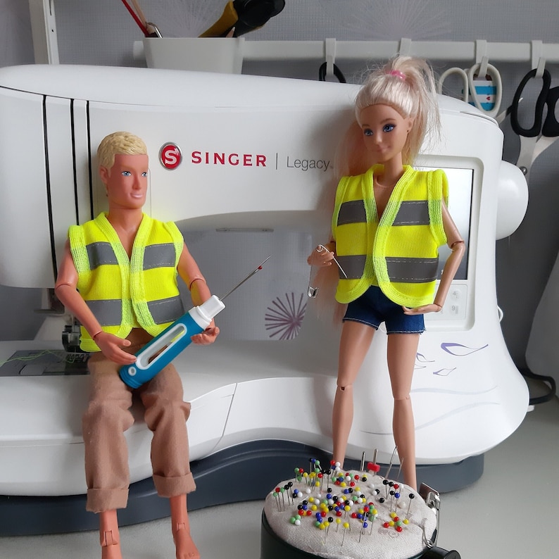 Puede incluir: Dos mu&ntilde;ecas con chalecos de seguridad amarillos y grises. Las mu&ntilde;ecas est&aacute;n de pie frente a una m&aacute;quina de coser blanca con el nombre de la marca "SINGER | Legacy" impreso en ella. Hay un taz&oacute;n de alfileres de colores delante de las mu&ntilde;ecas.