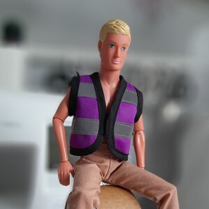 Puede incluir: Una mu&ntilde;eca con un chaleco de seguridad morado y gris con pantalones marrones.