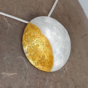 Könnte beinhalten: Eine kreisförmige Anhängerkette mit einem geteilten Design, das auf einer Seite eine texturierte goldfarbene Oberfläche und auf der anderen eine gebürstete silberfarbene Oberfläche aufweist. Der Anhänger hängt an einer dünnen, flexiblen silberfarbenen Schlangenkette.