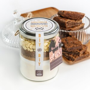May include: A glass jar filled with layered ingredients for a gingerbread brownie mix, labeled "DIY". The jar is topped with a metal lid and a tag. Several baked brownies are on a wooden board and in a glass dish.