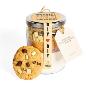 May include: A glass jar filled with ingredients for double chocolate chip cookies, including white chocolate chunks. A baked cookie with chocolate chips sits in front. The jar has a label that says "DOUBLE CHOCOLATE CHIP COOKIES" and a tag that says "DIY".