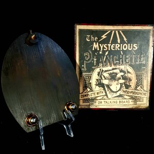 Puede incluir: Una planchette de madera vintage con un puntero de metal, utilizada para comunicarse con esp&iacute;ritus. La planchette est&aacute; sobre un soporte transparente. La caja dice "The Mysterious Planchette Complete with  Phsyco-Psychophone or Talking Board British Manufacture."