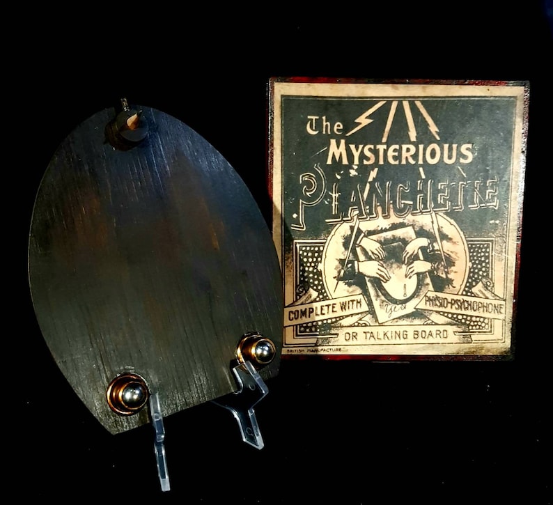 Puede incluir: Una planchette de madera vintage con un puntero de metal, utilizada para comunicarse con esp&iacute;ritus. La planchette est&aacute; sobre un soporte transparente. La caja dice "The Mysterious Planchette Complete with  Phsyco-Psychophone or Talking Board British Manufacture."