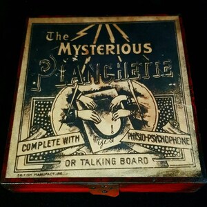 Puede incluir: Una caja de madera vintage con tapa roja y una etiqueta que dice "The Mysterious Planchette Complete with Physio-Psychophone or Talking Board." La etiqueta presenta una ilustraci&oacute;n en blanco y negro de manos que alcanzan una planchette.