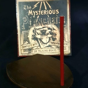 Puede incluir: Una planchette de madera vintage con un puntero rojo, utilizada para comunicarse con los esp&iacute;ritus. La planchette est&aacute; sobre una base de madera marr&oacute;n oscuro. La caja muestra una ilustraci&oacute;n de manos usando la planchette con el texto "The Mysterious Planchette Complete with Physio-Psychic or Talking Board British Manufacture".