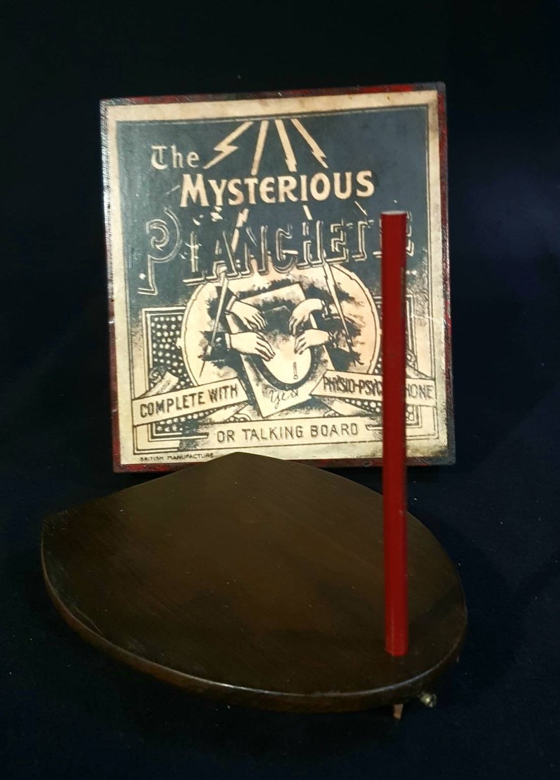Puede incluir: Una planchette de madera vintage con un puntero rojo, utilizada para comunicarse con los esp&iacute;ritus. La planchette est&aacute; sobre una base de madera marr&oacute;n oscuro. La caja muestra una ilustraci&oacute;n de manos usando la planchette con el texto "The Mysterious Planchette Complete with Physio-Psychic or Talking Board British Manufacture".