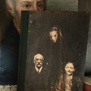 Op de afbeelding: Een vintage fotoalbum met een zwarte omslag en een rug met gouden letters. Het album is open op een pagina met een zwart-witfoto van drie mensen. De foto toont een man met een snor en een vrouw met lang haar, beiden zittend, en een jongere persoon die achter hen staat.