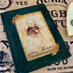 Puede incluir: Un libro de tapa dura verde con una foto de un hombre en la portada. El libro se titula "Allan Kardec" y está sobre una superficie de madera con una tabla Ouija en el fondo.
