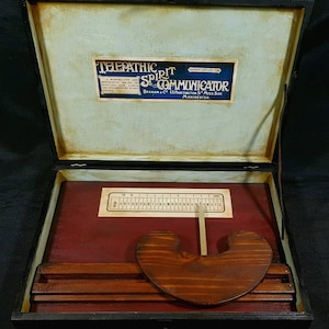May include: A wooden box with a red interior and a sliding wooden pointer. The box contains a wooden alphabet board and a label that reads "Telepathic Spirit Communicator, Braham & Co. 13 Hartington St. Moss Side, Manchester."