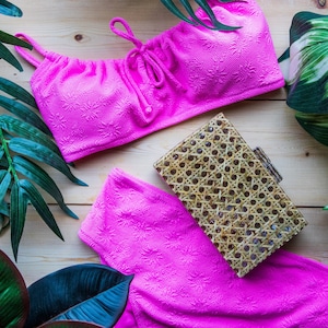 Może przedstawiać: Różowy top i dół bikini z plecioną wiklinową kopertówką. Bikini ma kwiatowy wzór, a kopertówka ma prostokątny kształt.