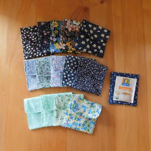 May include: A collection of fabric tea bag holders in various floral patterns, including white daisies on navy, pink and yellow flowers, and blue floral prints. One holder contains a tea bag with the text "Organics Chamomile".