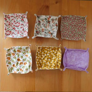 May include: Six fabric trays with floral and paisley patterns. The trays are square with rounded corners and small handles. The patterns include red roses, orange and white flowers, small red flowers, green and orange paisley, yellow flowers, and a purple tie-dye.