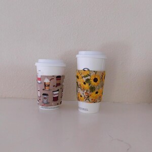 May include: Two white disposable coffee cups with fabric sleeves. The smaller cup features a beige sleeve with a pattern of various coffee drinks and beans. The larger cup has a sleeve decorated with a vibrant yellow sunflower print.
