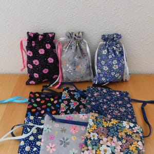 May include: A collection of drawstring bags in various floral patterns. The bags feature a range of colors, including black with pink flowers, gray with white flowers, and blue with white flowers. Each bag has a matching ribbon closure.