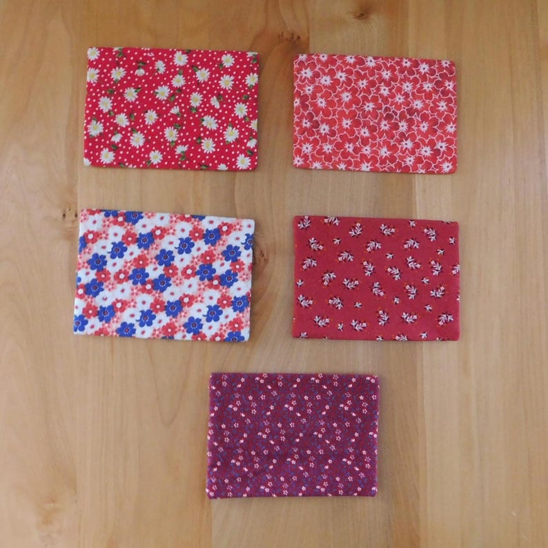 May include: Five rectangular fabric coasters featuring various floral patterns. The top left is red with white daisies, the top right is red with white flowers, the middle left is white with blue and red flowers, the middle right is red with small white sprigs, and the bottom is purple with tiny white flowers.