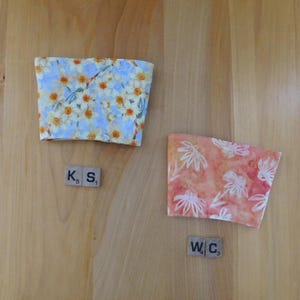 May include: Two fabric cup sleeves. One sleeve features a light blue background with yellow and white daffodil flowers. The other sleeve has a coral and orange background with white floral outlines. Two small square tiles labeled K5 and S1 are near the first sleeve, and tiles labeled W4 and C3 are near the second.