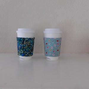 May include: Two white disposable coffee cups with fabric sleeves. The left sleeve features a dark blue and green leaf pattern. The right sleeve is light blue with a repeating pattern of small red, yellow, and white flowers on green stems.