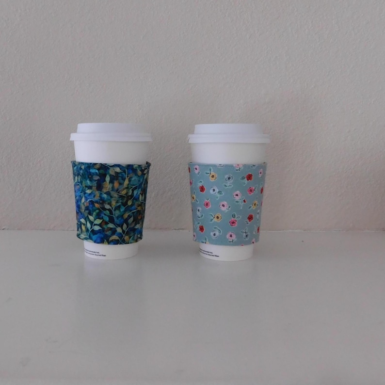 May include: Two white disposable coffee cups with fabric sleeves. The left sleeve features a dark blue and green leaf pattern. The right sleeve is light blue with a repeating pattern of small red, yellow, and white flowers on green stems.