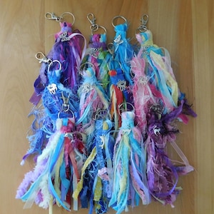 May include: A collection of multi-colored fabric tassel keychains, each featuring a silver-toned metal lobster clasp and a small silver-toned charm. The tassels are composed of various ribbons, lace, and yarn strips in shades of blue, purple, pink, and yellow.