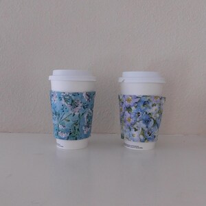 May include: Two white disposable coffee cups with lids, each featuring a fabric sleeve. The left sleeve has a light blue background with small white and pink floral patterns. The right sleeve displays a dense pattern of blue and purple flowers.